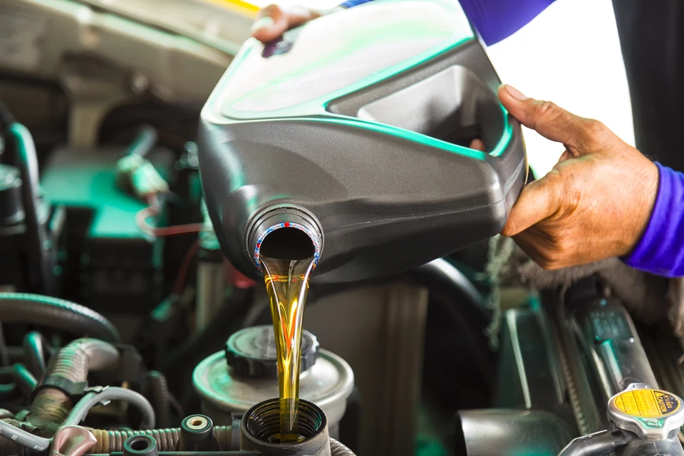 Mécanicien automobile versant de l'huile moteur dorée depuis un bidon noir dans le compartiment moteur d'un véhicule lors d'une vidange dans un garage professionnel