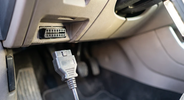 Technicien automobile en bleu de travail utilisant un dispositif de diagnostic électronique branché au système OBD d'un véhicule avec capot ouvert. Le diagnostic affiche des codes d'erreur sur écran de test. L'arrière-plan montre l'intérieur d'un atelier garage moderne avec outils de diagnostic, établi et équipements professionnels. Prise de vue détaillée montrant la précision et la fiabilité du diagnostic électronique automobile.